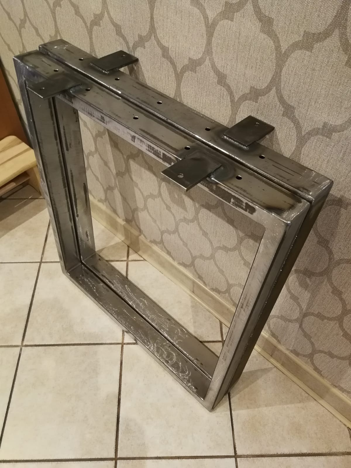 Rama stołu z profili stalowych, spawana, z widocznymi punktami mocowania blatu, surowy metal, tło: płytki podłogowe i wzorzysta tapeta.