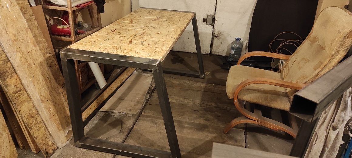 Stół roboczy z metalową ramą i blatem z płyty OSB w warsztacie, widoczne elementy spawane i surowe wykończenie.