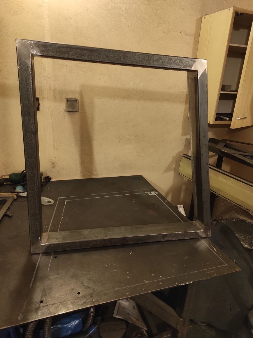 Surowa, kwadratowa rama z metalowych profili, prawdopodobnie w trakcie realizacji konstrukcji stalowej, widoczna na warsztatowym stole z zaznaczonym obrysem elementu do dospawania.