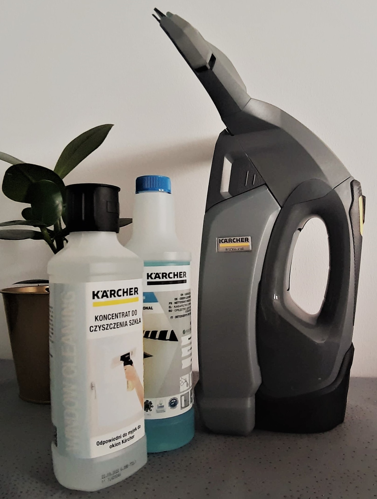 Zestaw do mycia okien Kärcher: elektryczna myjka do okien, koncentrat do czyszczenia szkła i płyn do mycia okien, ustawione obok doniczki z rośliną na blacie.