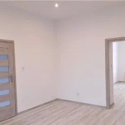 Pusty, wyremontowany pokój z panelami podłogowymi i dwiema parami drzwi: jedne z przeszkleniami, drugie otwarte na pokój z grzejnikiem pod oknem.