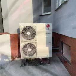 Biała pompa ciepła LG Therma V z dwoma wentylatorami, zamontowana na zewnątrz budynku z szarą elewacją i czerwoną cegłą, oświetlona promieniami słońca.