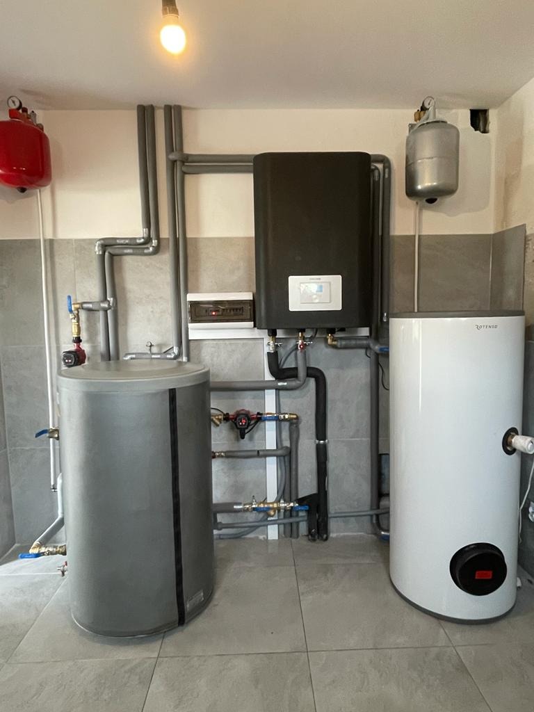 Kompletna instalacja grzewcza w kotłowni: dwa zbiorniki na wodę (szary i biały), wiszący czarny piec, rury izolowane termicznie, czerwony zbiornik wyrównawczy, szara płytki na podłodze i ścianach.