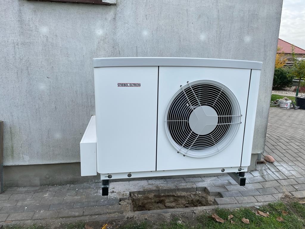Biała jednostka zewnętrzna z wentylatorem marki Stiebel Eltron zamontowana na czarnych wspornikach, widoczny wykop w kostce brukowej obok urządzenia.