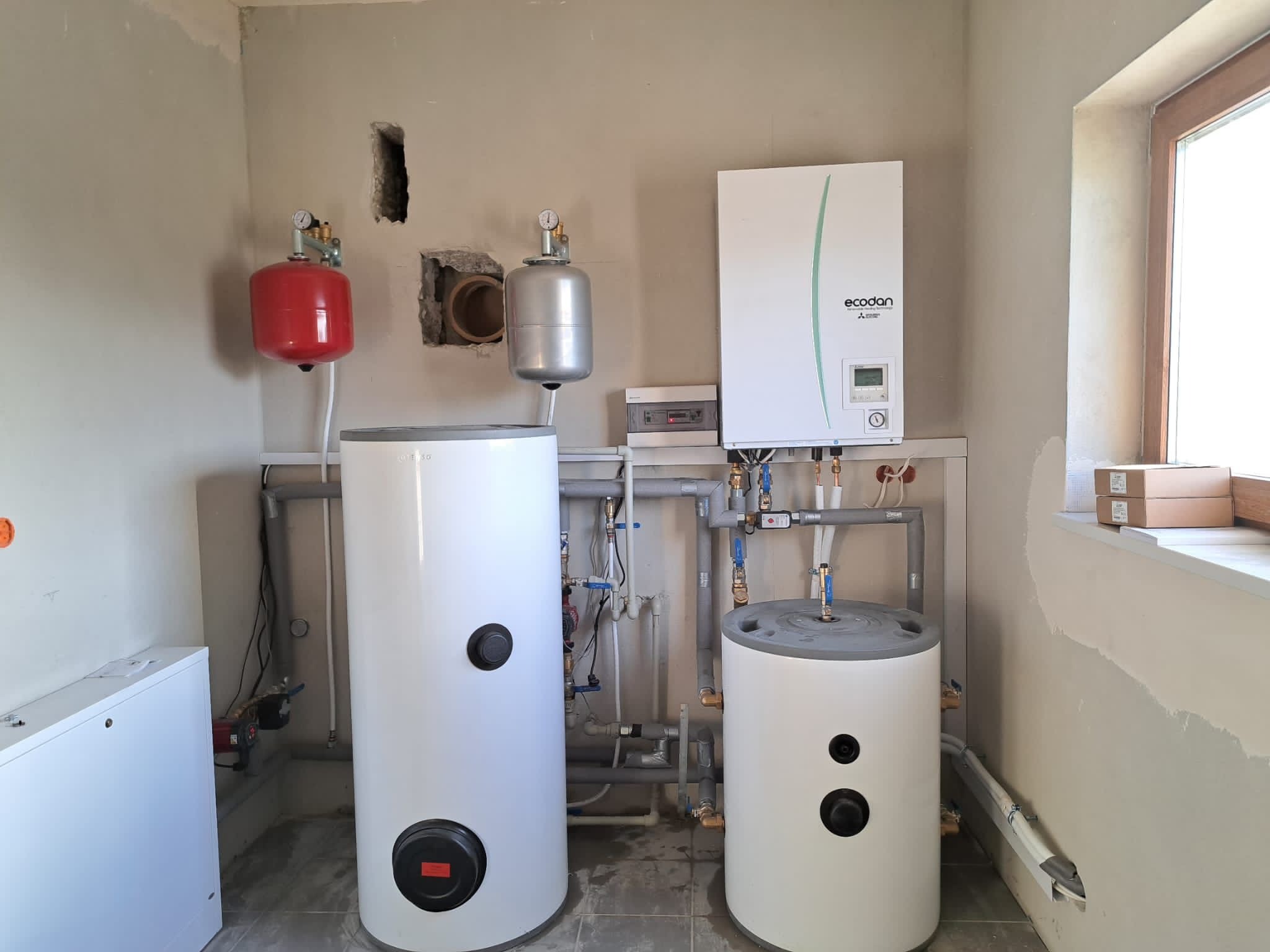Instalacja grzewcza w surowym pomieszczeniu: dwa białe zbiorniki, czerwony i srebrny zbiornik wyrównawczy, biały panel sterowania marki Ecodan, rury i okablowanie na tle szarej, nieotynkowanej ściany.