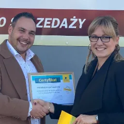 Wręczenie certyfikatu 'Odpowiedzialnego Pracodawcy' przez przedstawicielkę firmy ubezpieczeniowej Aviva, uścisk dłoni, uśmiechnięci ludzie na tle ściany z napisem 'Biuro Sprzedaży Fabryka Smaku'.