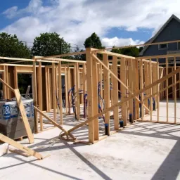 Wznoszenie drewnianej konstrukcji budynku z widocznymi instalacjami hydraulicznymi, bloczkami betonowymi i elementami deskowania na betonowej płycie fundamentowej, z domem w tle.