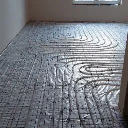 Białe rury ogrzewania podłogowego ułożone w pętle na folii z nadrukowaną siatką i logo Altech, widok z góry w surowym wnętrzu z oknem.