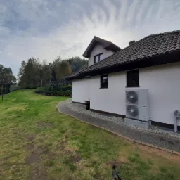 Biały dom jednorodzinny z szarą dachówką, na ścianie zamontowana dwuwentylatorowa jednostka zewnętrzna, obok chodnik z kostki brukowej, trawnik i rząd tuj.