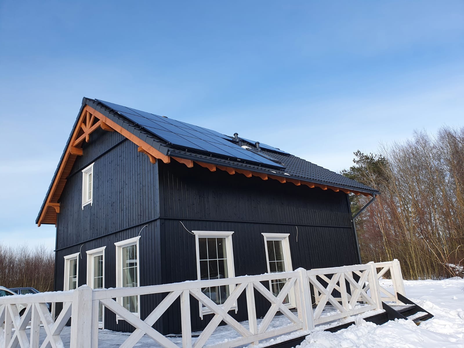 Czarny dom z panelami słonecznymi na dachu, widoczny z perspektywy ogrodu pokrytego śniegiem i białym płotem.