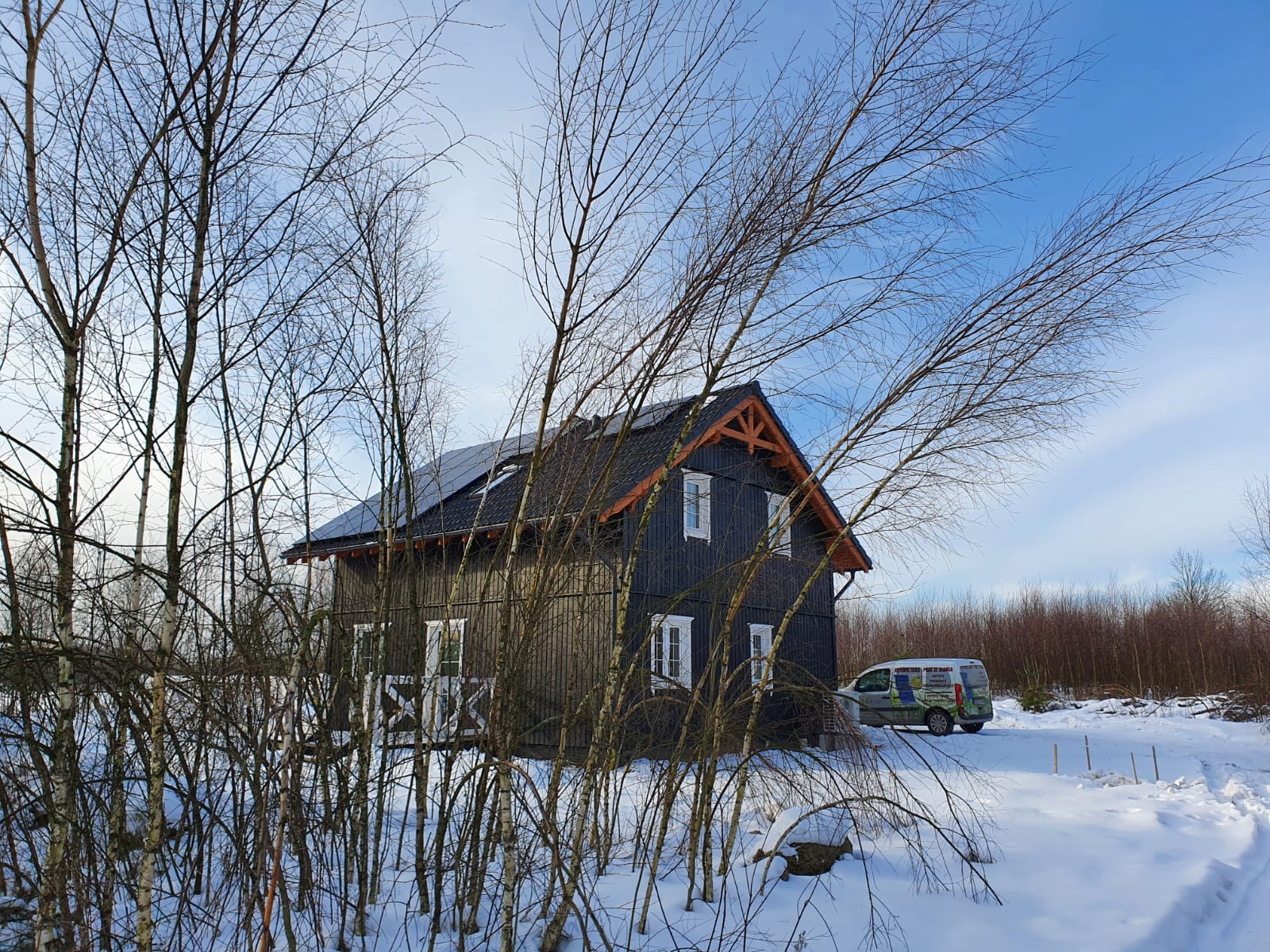 Ciemny, drewniany dom z panelami słonecznymi na dachu, otoczony ośnieżonym krajobrazem i młodymi brzózkami bez liści, z samochodem serwisowym w tle.