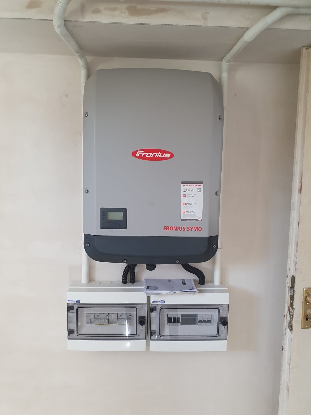 Szary inwerter Fronius SYMO zamontowany na ścianie nad dwoma skrzynkami bezpiecznikowymi, z widocznymi przewodami i dokumentacją techniczną na górze skrzynek.