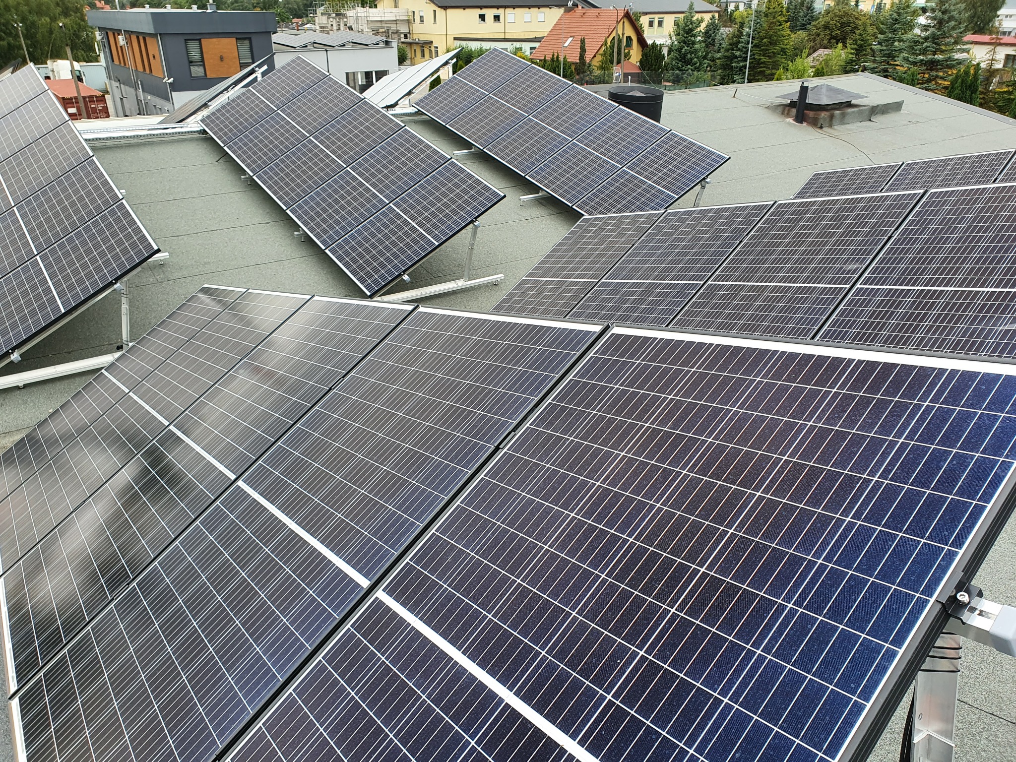 Instalacja paneli słonecznych na płaskim dachu budynku, widok z góry, równomiernie rozłożone moduły z aluminiowymi konstrukcjami wsporczymi, w tle zabudowa mieszkalna i drzewa.