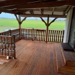 Zadaszony taras z drewnianą podłogą i ozdobną balustradą, widok na zielone pole w tle, filiżanki na balustradzie.