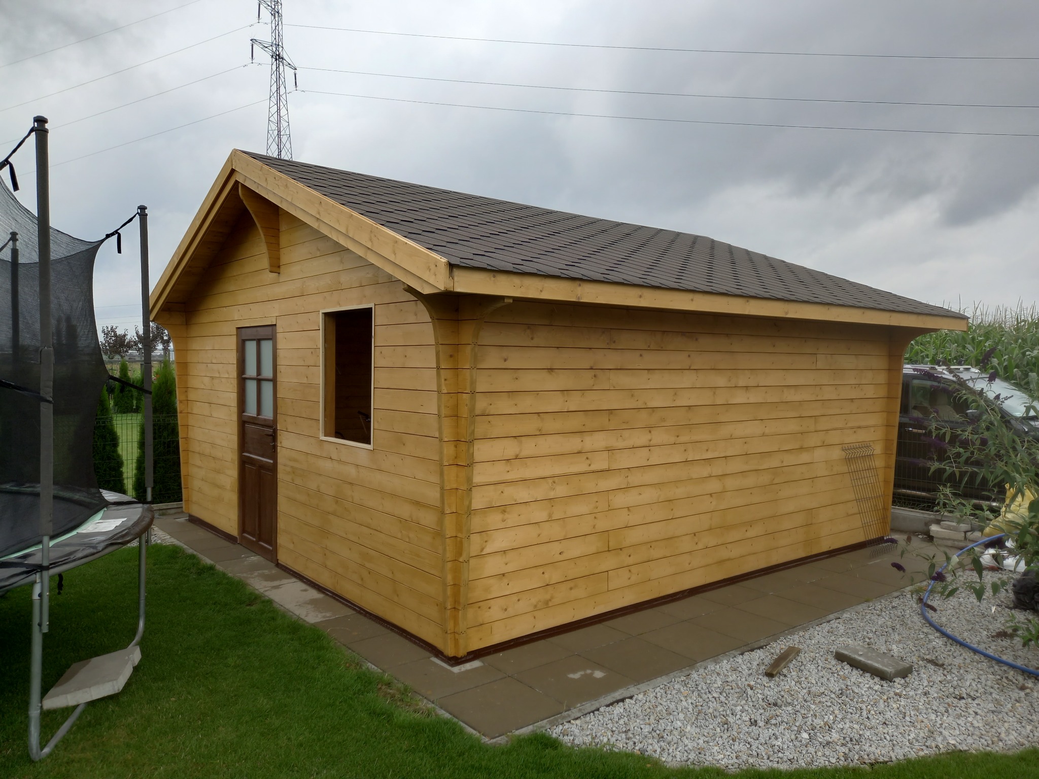 Gotowa drewniana altana z brązowym dachem bitumicznym, brązowymi drzwiami i oknem bez szyby, na betonowej wylewce, obok trampoliny, na tle pochmurnego nieba i słupów wysokiego napięcia.