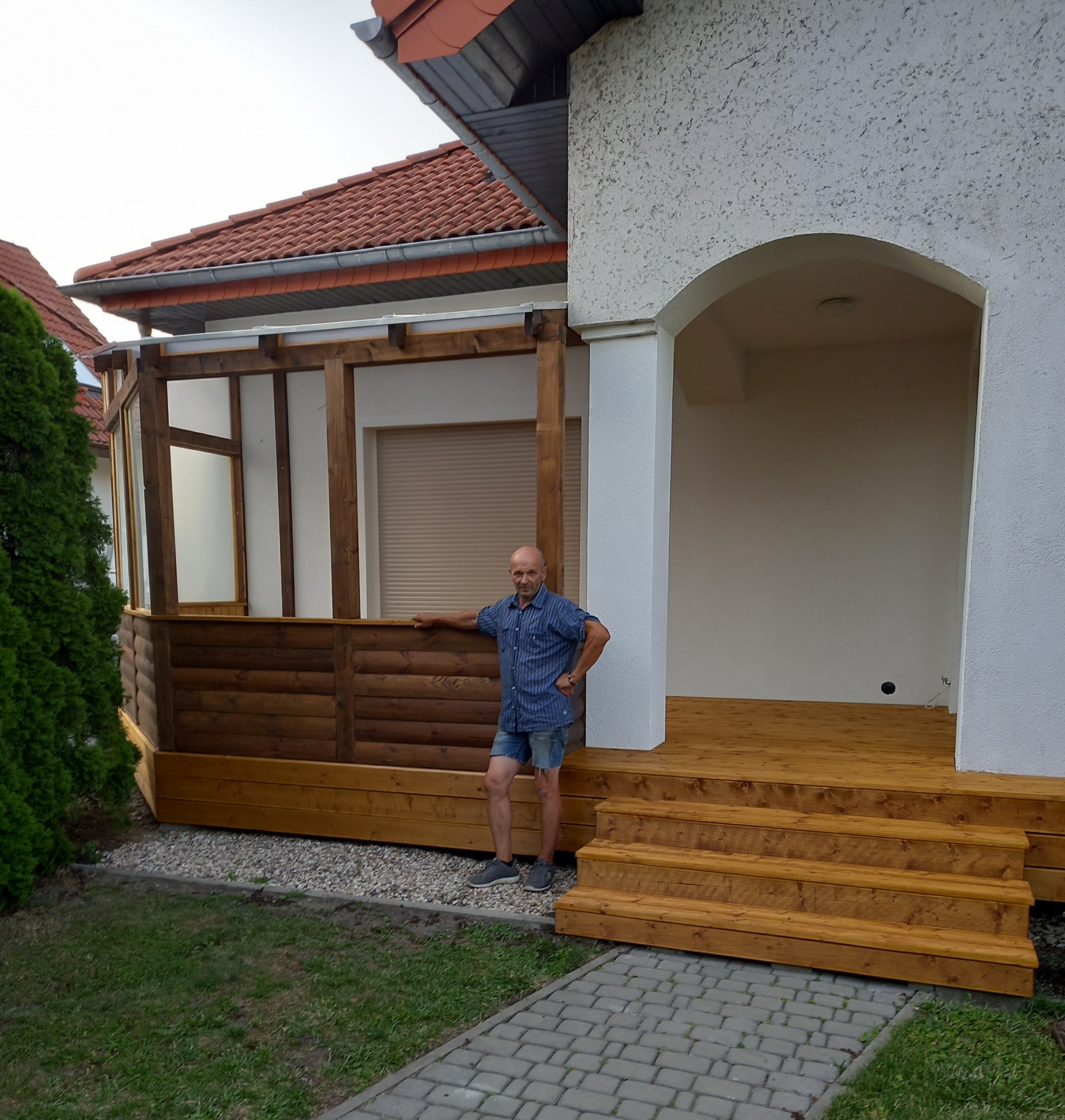 Mężczyzna w niebieskiej koszuli i dżinsowych spodenkach stoi na drewnianym tarasie przylegającym do domu z białymi ścianami i czerwonym dachem, obok częściowo zabudowanej drewnianej konstrukcji.