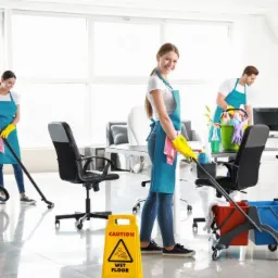 Zespół sprzątający w jasnym biurze: uśmiechnięta kobieta z mopem na pierwszym planie, odkurzacz i znak ostrzegawczy 'mokra podłoga' w tle, pracownicy w niebieskich fartuchach i żółtych rękawicach.