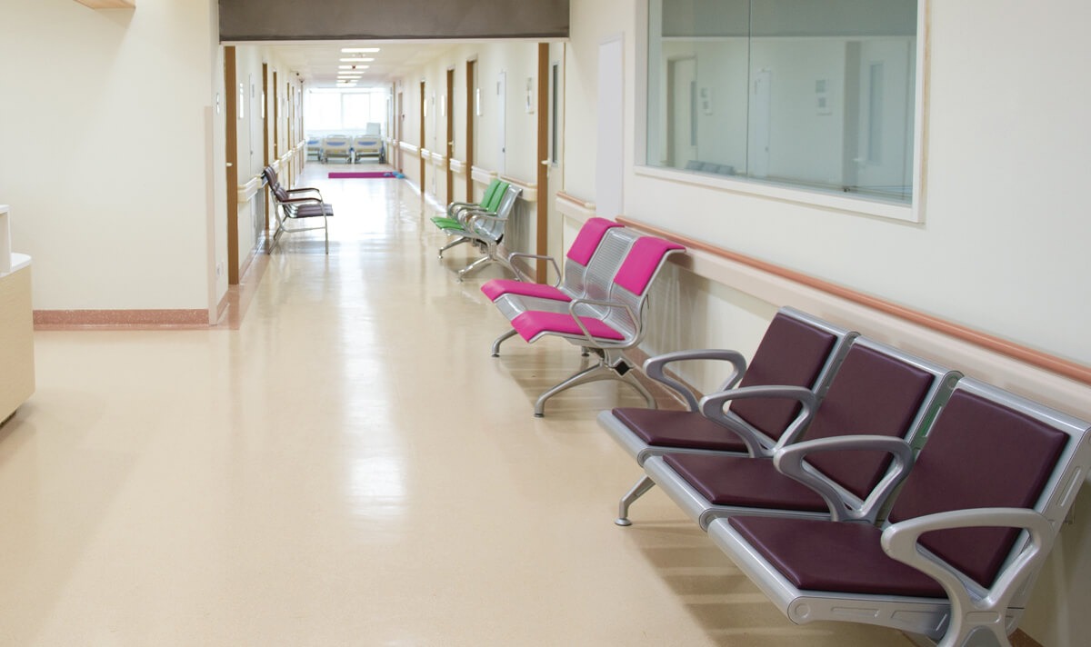 Długi, jasny korytarz w szpitalu lub biurze z rzędami metalowych krzeseł z kolorowymi siedziskami i oparciami, błyszcząca podłoga, widok na salę z łóżkami na końcu korytarza.