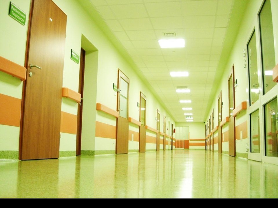 Długi, jasny korytarz w szpitalu lub klinice z błyszczącą, świeżo umytą podłogą i rzędami zamkniętych drzwi po obu stronach.