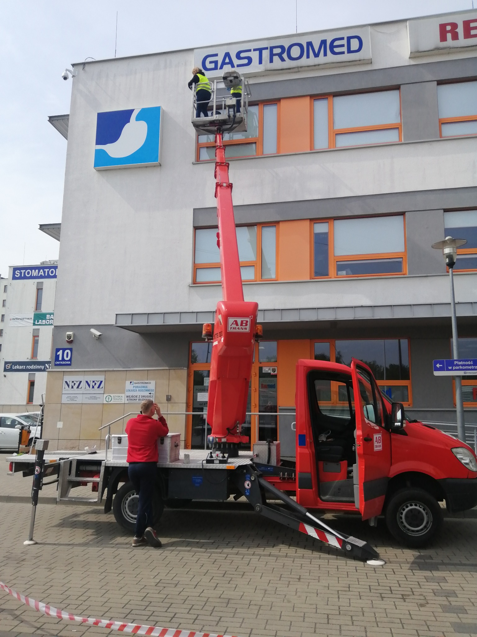 Pracownicy na podnośniku koszowym montują logo Gastromed na elewacji budynku, widoczny czerwony samochód z otwartymi drzwiami i zabezpieczenia na chodniku.