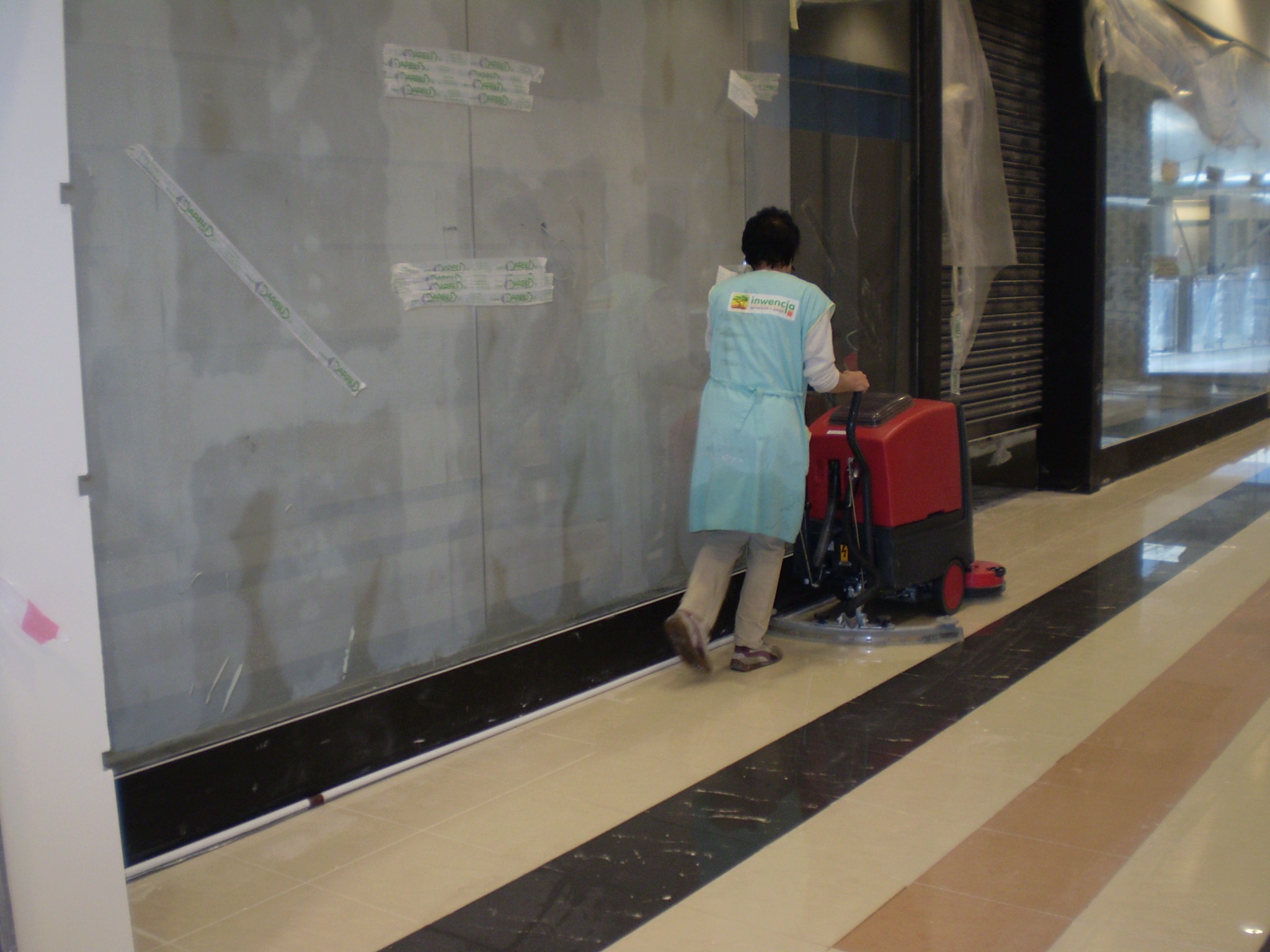 Pracownik w niebieskim fartuchu z logo firmy obsługuje czerwoną maszynę do czyszczenia podłóg w pasażu handlowym, widoczne ślady po remoncie na szklanej ściance.