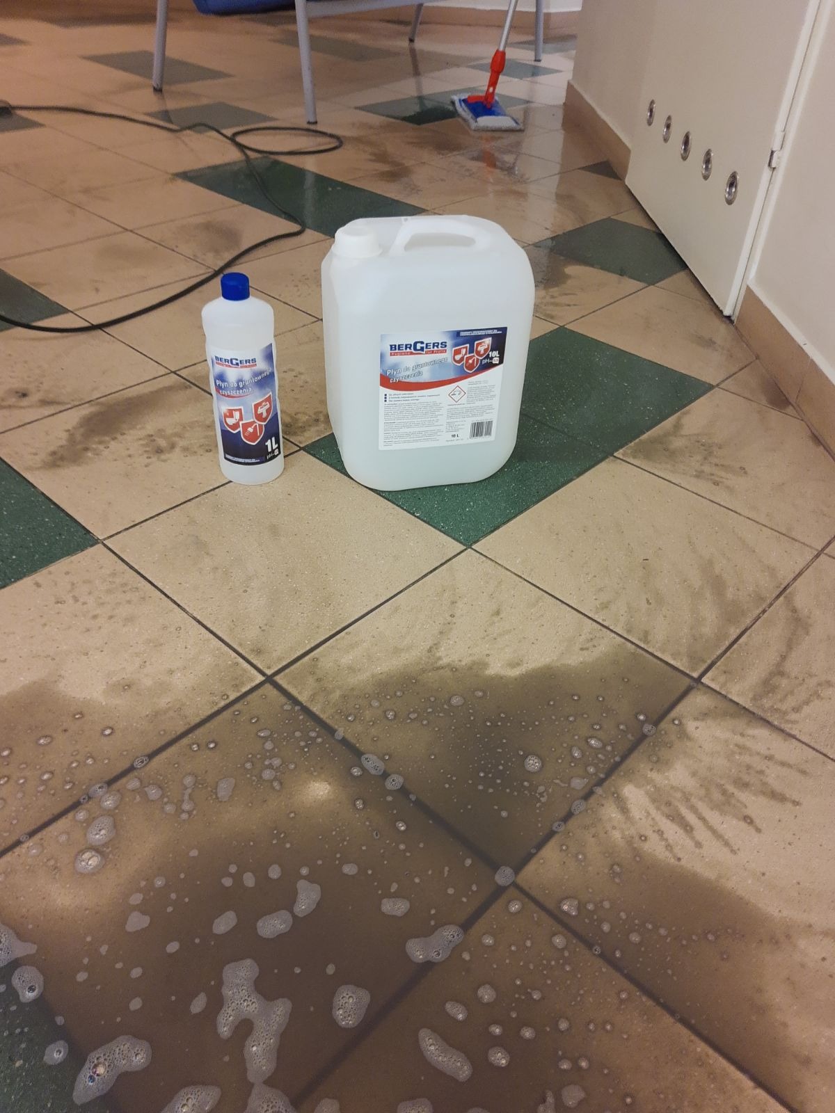 Płytki podłogowe w trakcie czyszczenia, widoczne rozlane środki czystości marki Bergers w opakowaniach 1L i 10L oraz mop w tle.