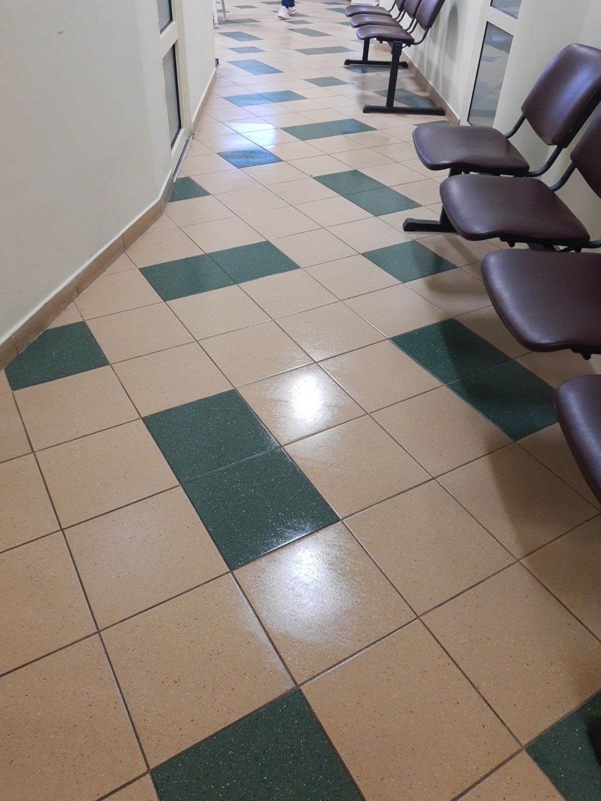 Błyszcząca, świeżo umyta podłoga z płytek w odcieniach beżu i zieleni ułożonych w szachownicę w korytarzu z krzesłami.