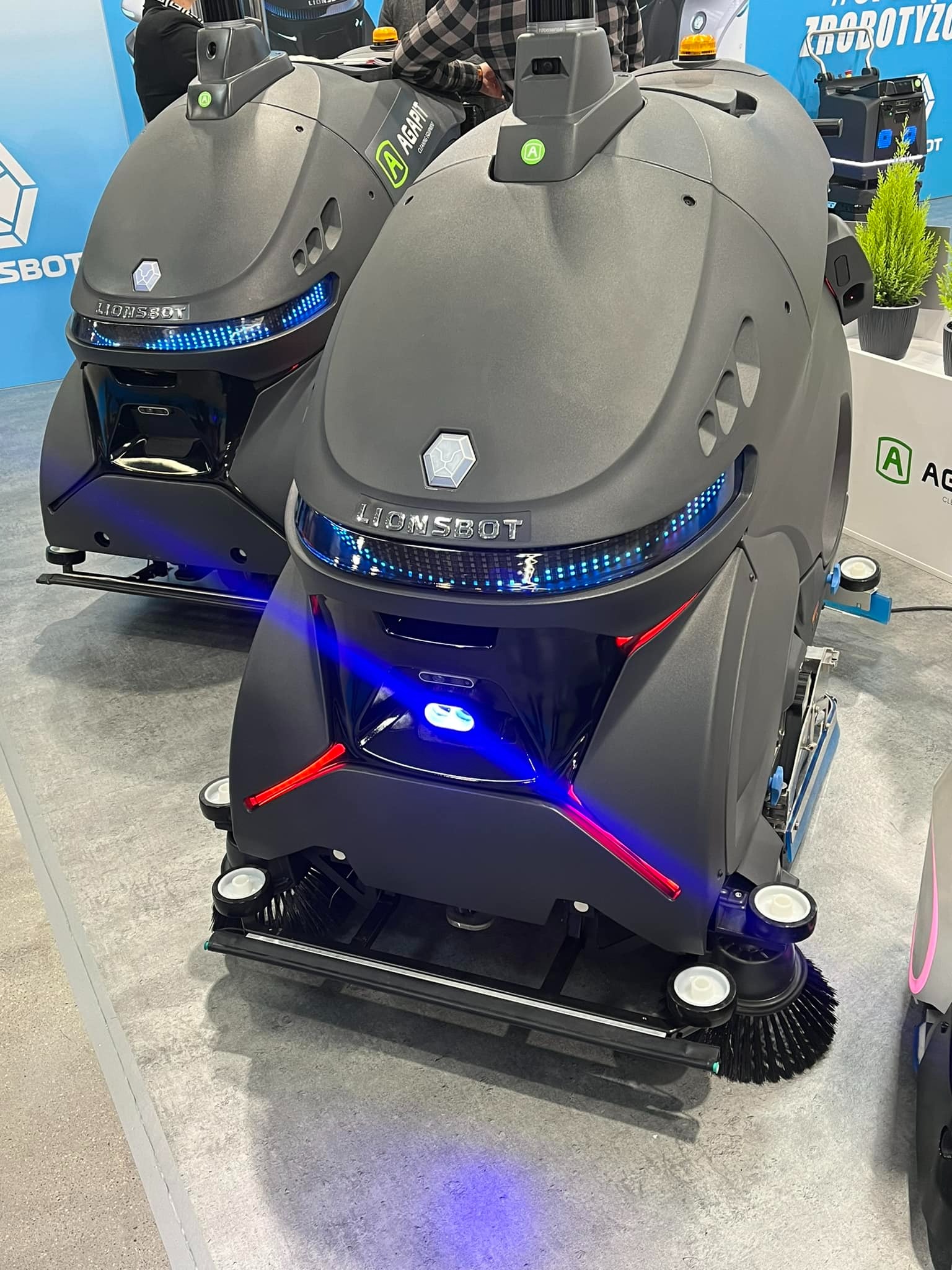 Dwa roboty czyszczące Lionsbot z niebieskim i czerwonym oświetleniem LED, widoczne szczotki i logo Agapit na tle ekspozycji targowej.