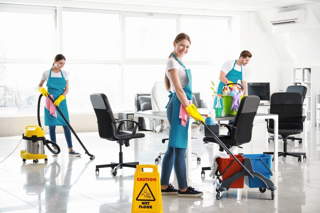 Zespół sprzątający w jasnym biurze: uśmiechnięta kobieta z mopem na pierwszym planie, odkurzacz i znak ostrzegawczy 'mokra podłoga' w tle, pracownicy w niebieskich fartuchach i żółtych rękawicach.