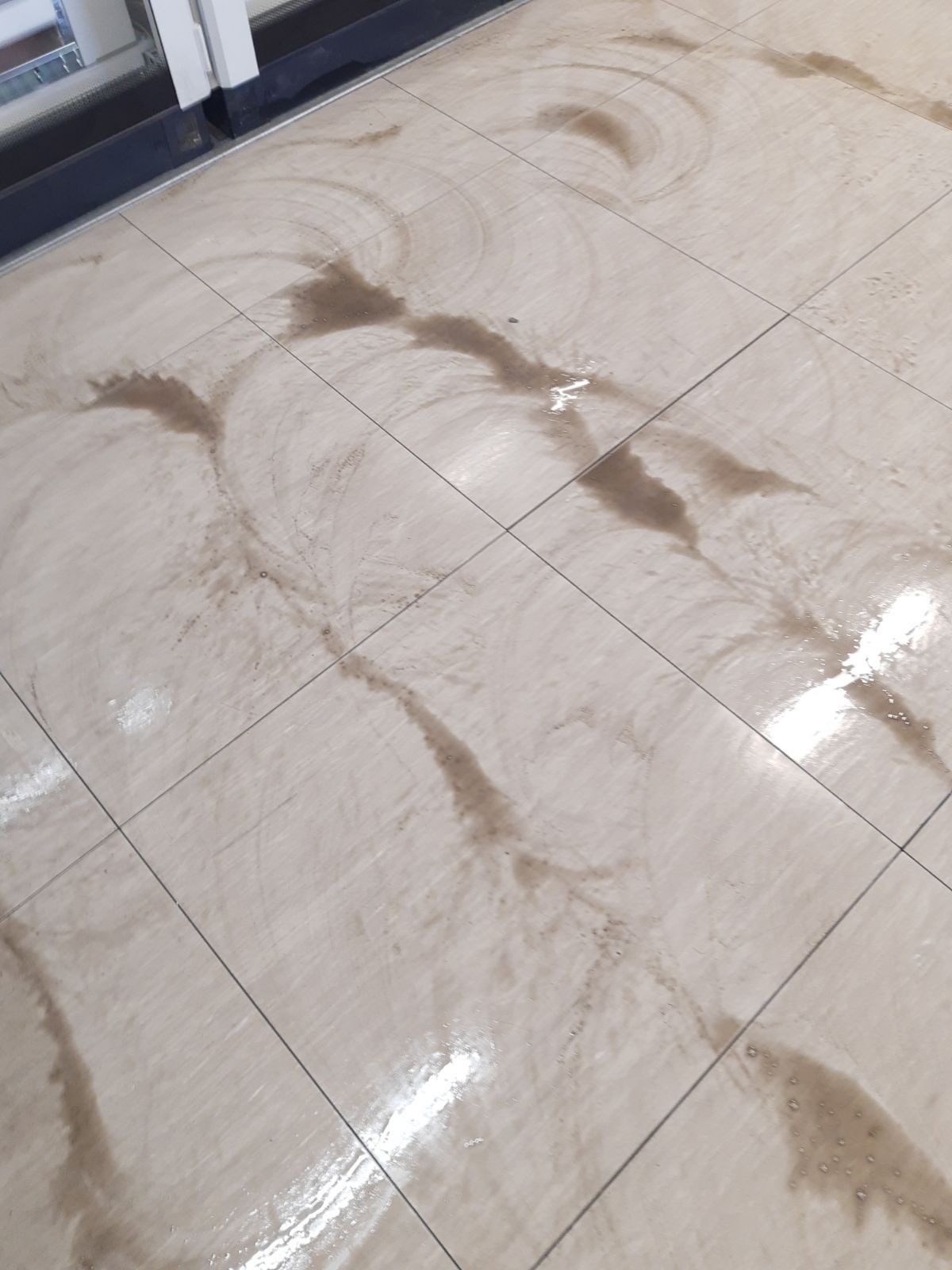 Beżowe płytki podłogowe z widocznymi śladami brudnej wody i zaciekami po sprzątaniu, w pobliżu drzwi wejściowych.
