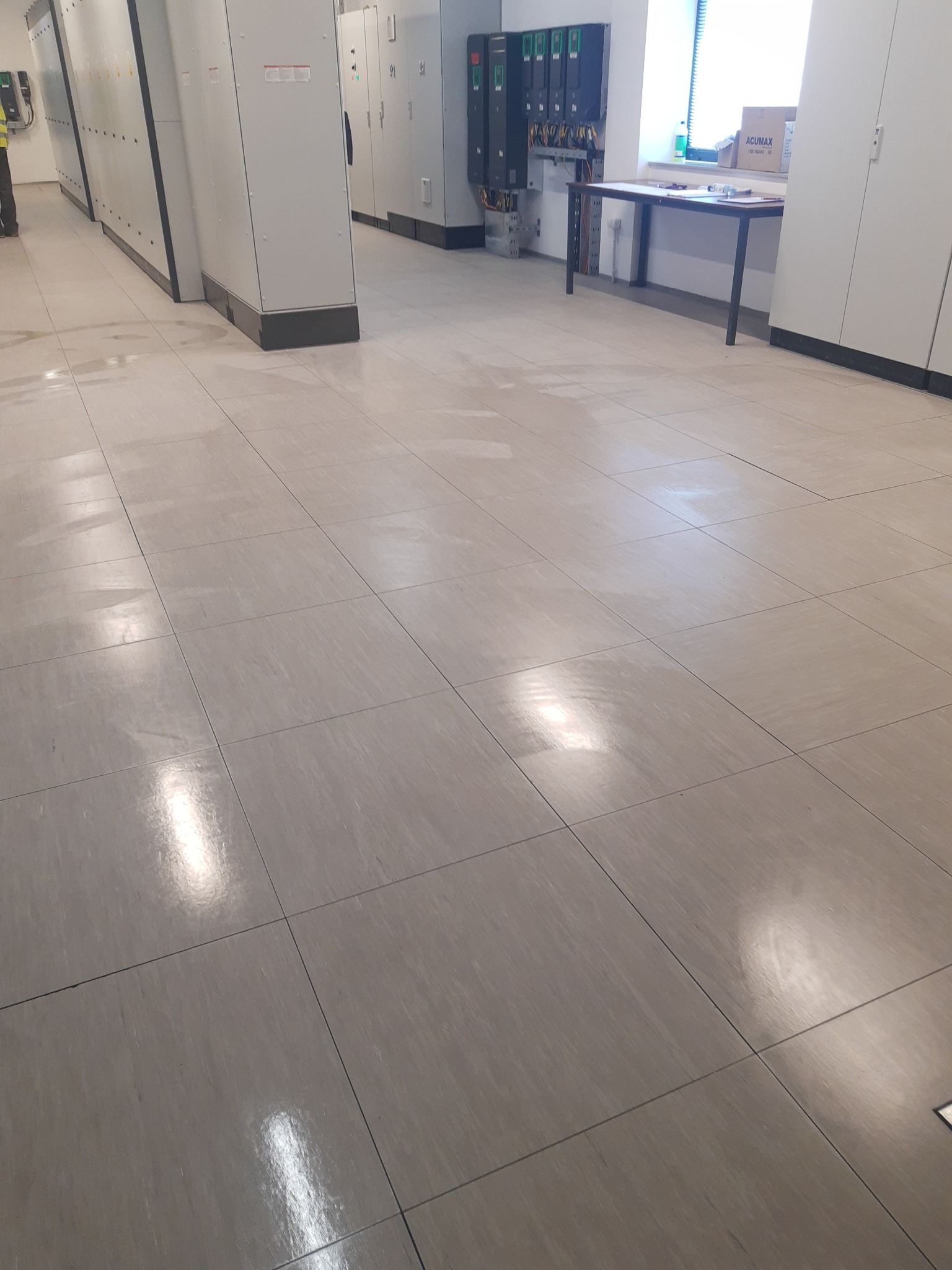 Wyślizgana, świeżo umyta podłoga z płytek w hali przemysłowej, z rzędami szaf elektrycznych w tle i stołem przy oknie.