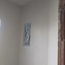 Świeżo otynkowana ściana w rogu pomieszczenia, z widoczną puszką instalacyjną z przewodami elektrycznymi i fragmentem ściany z odsłoniętą cegłą.