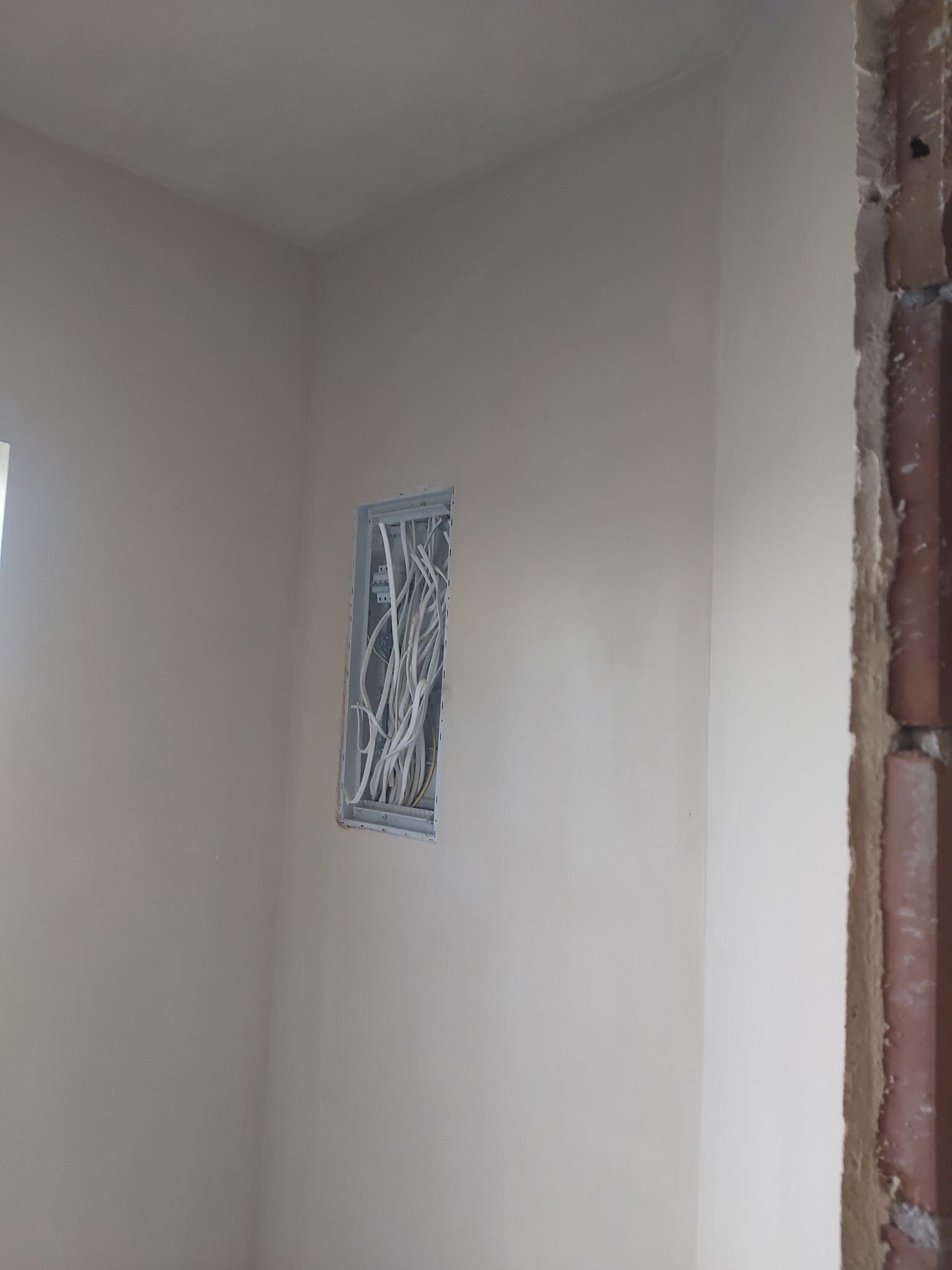 Świeżo otynkowana ściana w rogu pomieszczenia, z widoczną puszką instalacyjną z przewodami elektrycznymi i fragmentem ściany z odsłoniętą cegłą.