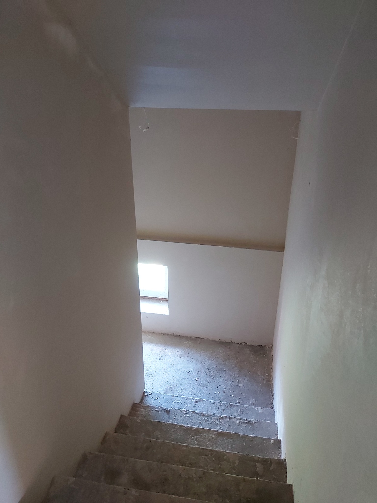 Klatka schodowa w surowym stanie po pracach budowlanych, ściany pokryte świeżym tynkiem, widoczne schody i fragment okna w tle.