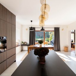 Nowoczesna kuchnia z jadalnią: ciemne drewniane szafki, czarny blat wyspy, okrągłe lustro i widok na ogród przez okno. Minimalistyczny design z akcentami natury.