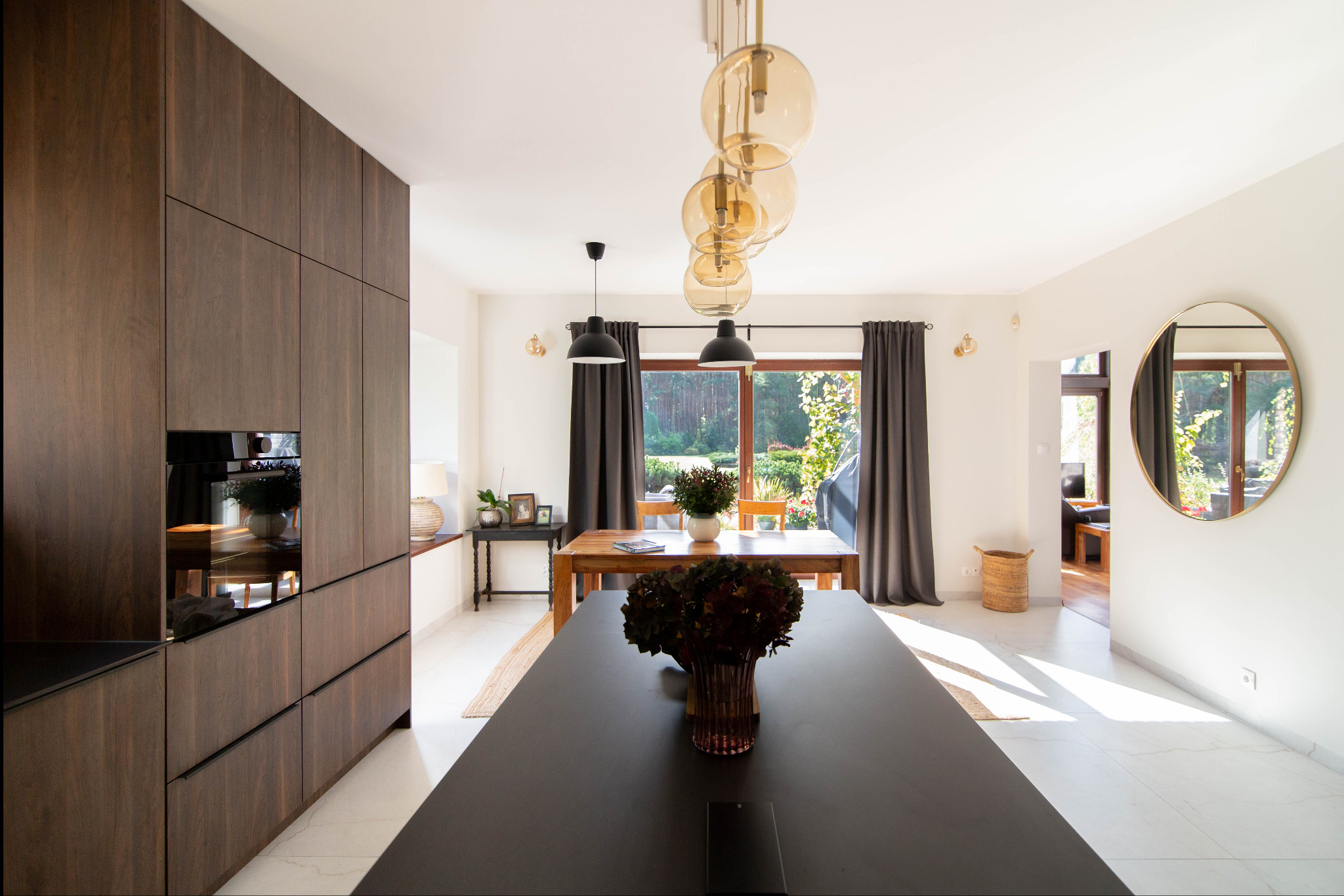 Nowoczesna kuchnia z jadalnią: ciemne drewniane szafki, czarny blat wyspy, okrągłe lustro i widok na ogród przez okno. Minimalistyczny design z akcentami natury.