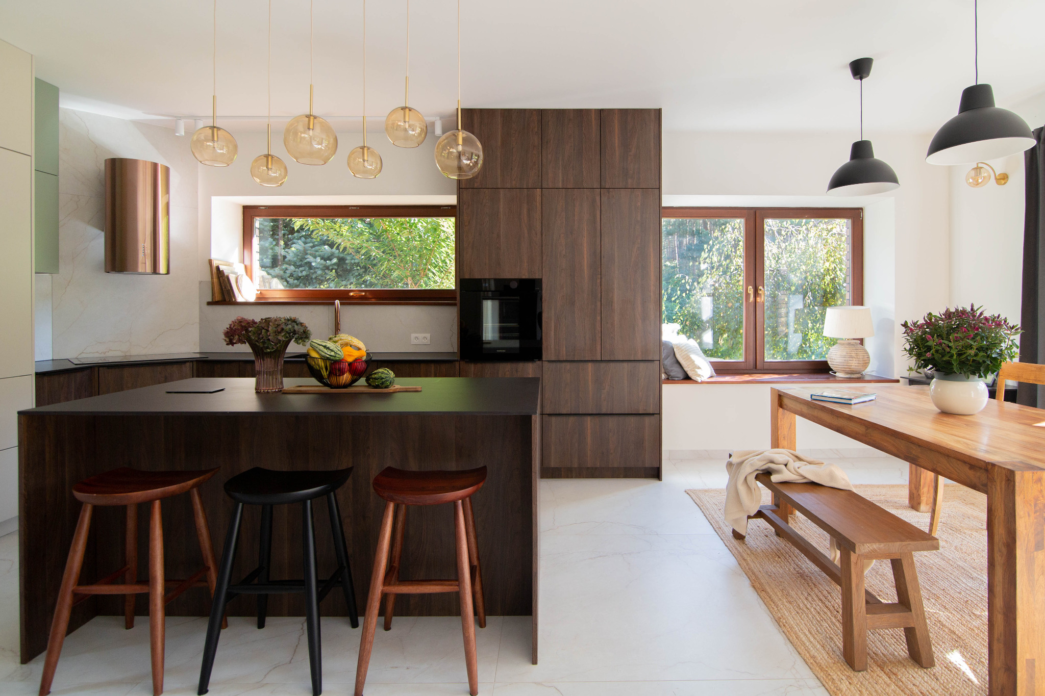 Nowoczesna kuchnia z jadalnią: ciemne drewno, wyspa z hokerami, okno z widokiem na zieleń, lampy wiszące. Minimalistyczny design, naturalne światło, marmurowe płytki.