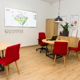Jasne biuro z drewnianą podłogą, czerwonymi krzesłami, okrągłymi stołami i tablicą interaktywną z wizualizacją domu oraz logo firmy ERGO HESTIA.