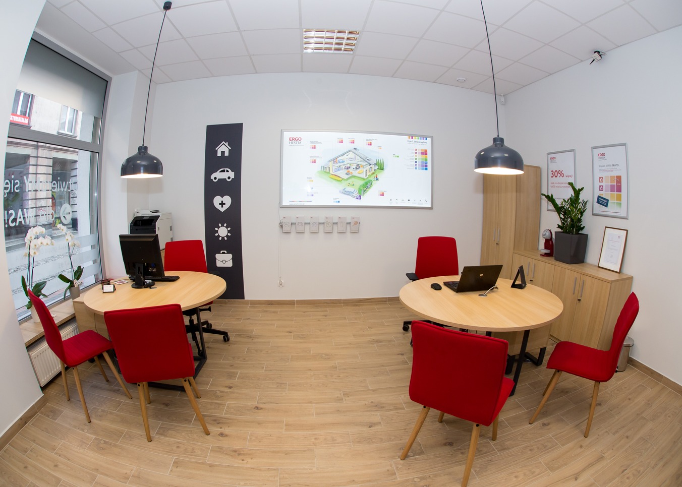 Jasne biuro ubezpieczeniowe z dwoma okrągłymi stołami, czerwonymi krzesłami, tablicą interaktywną z rysunkiem domu i samochodu, oraz ścianą z ikonami: domu, auta, serca, słońca i teczki.