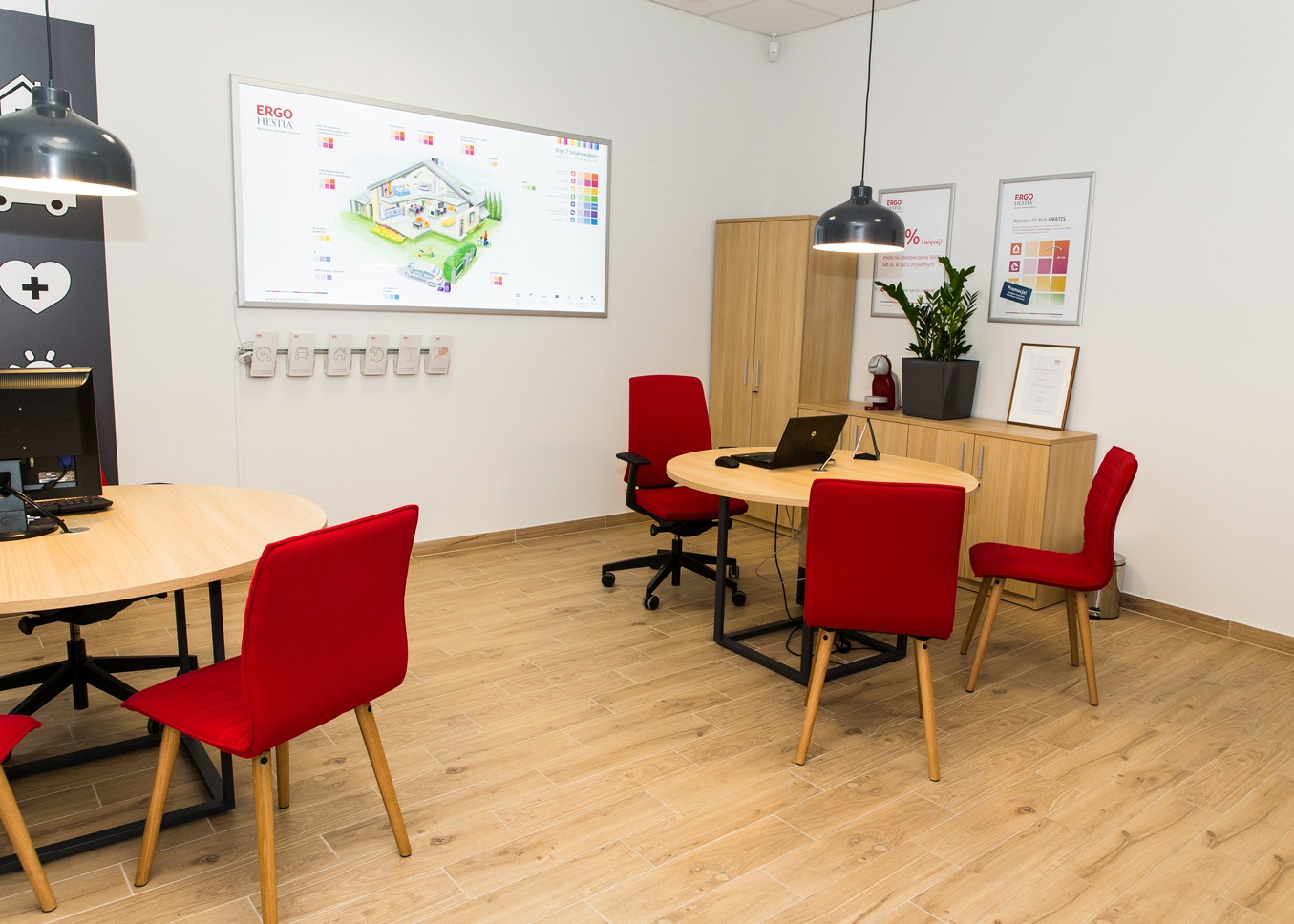 Jasne biuro z drewnianą podłogą, czerwonymi krzesłami, okrągłymi stołami i tablicą interaktywną z wizualizacją domu oraz logo firmy ERGO HESTIA.