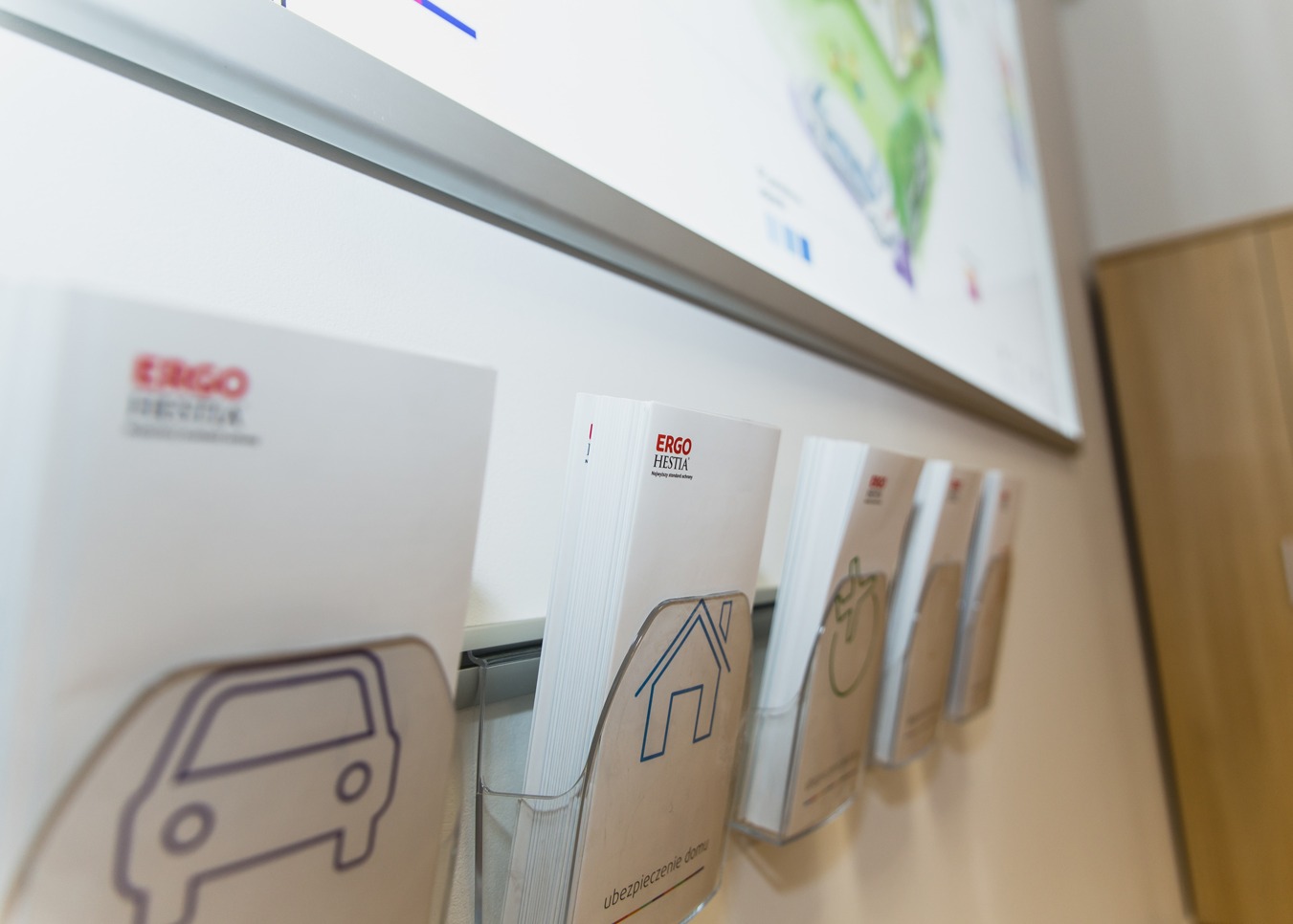 Ulotki informacyjne ERGO Hestia w przezroczystych kieszeniach na ścianie, z ikonami samochodu, domu i jabłka, sugerujące różne rodzaje ubezpieczeń, podświetlone przez światło z góry.