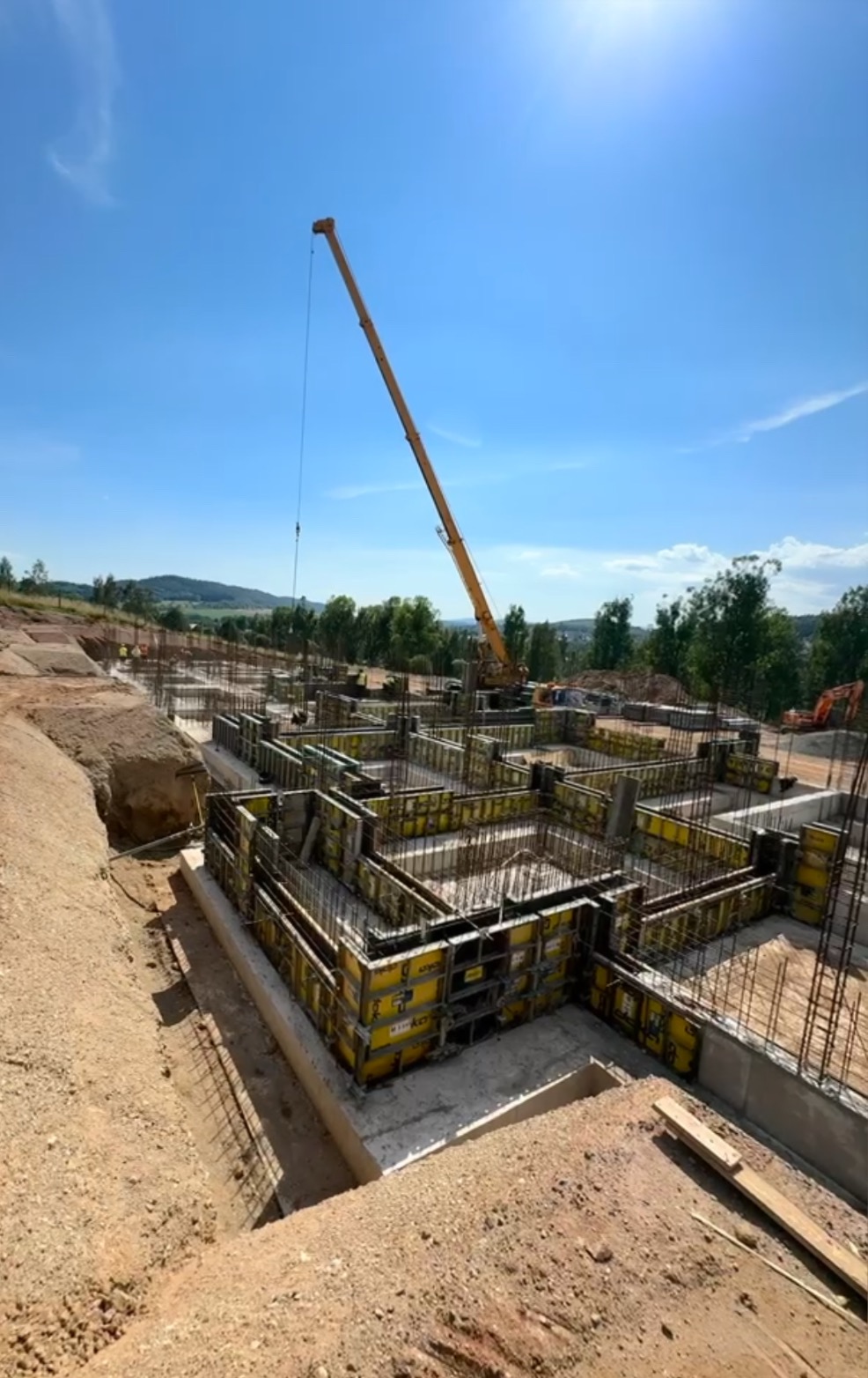 Żółte szalunki fundamentów zbrojonych stalą, widok z góry, na tle krajobrazu i nieba. Na budowie stoi żuraw. Słoneczny dzień.