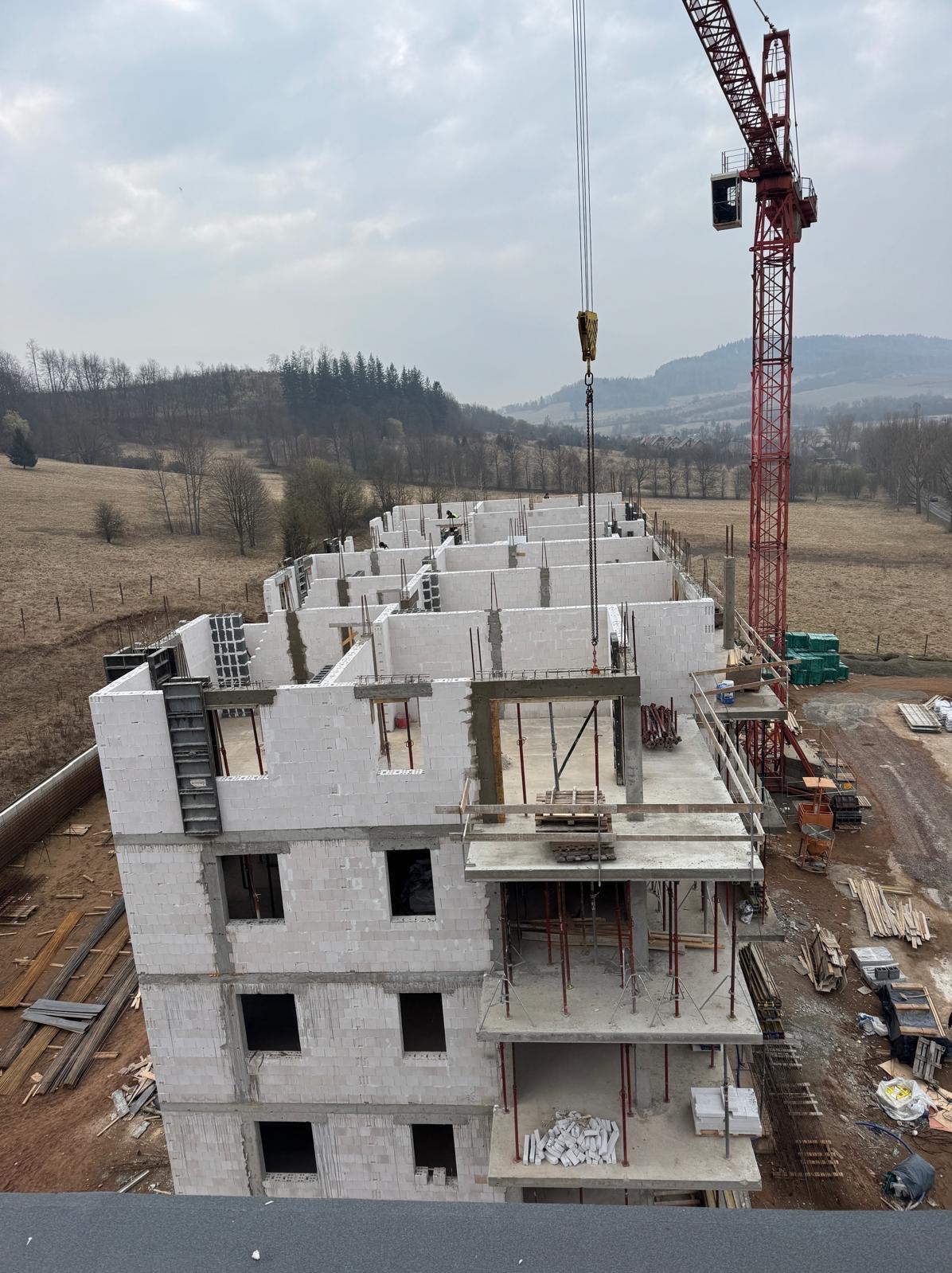 Wznoszenie wielopiętrowego budynku z białych bloczków, widok z góry. Na budowie czerwony dźwig i materiały budowlane. W tle zalesione wzgórza.