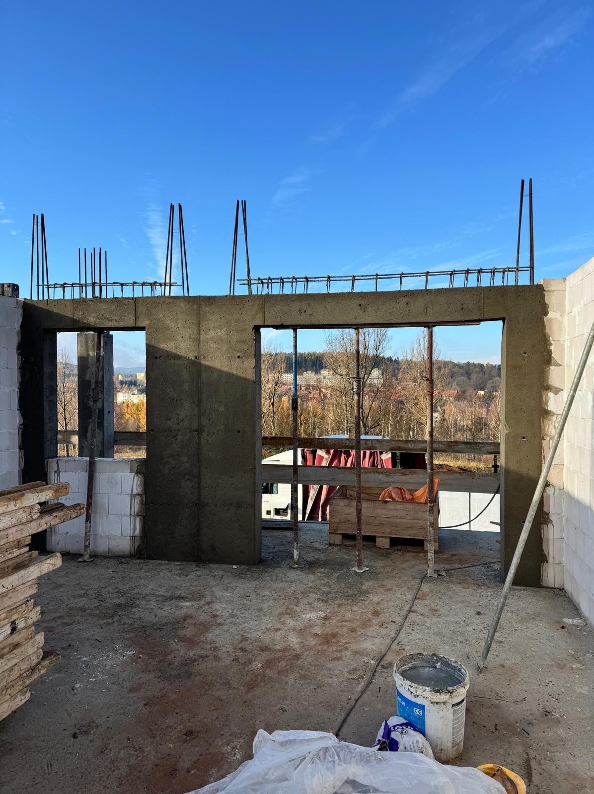 Surowy stan budynku w trakcie budowy: ściany z betonu komórkowego, zbrojenia wystające ponad wieniec, widok na krajobraz przez otwory okienne i drzwiowe.