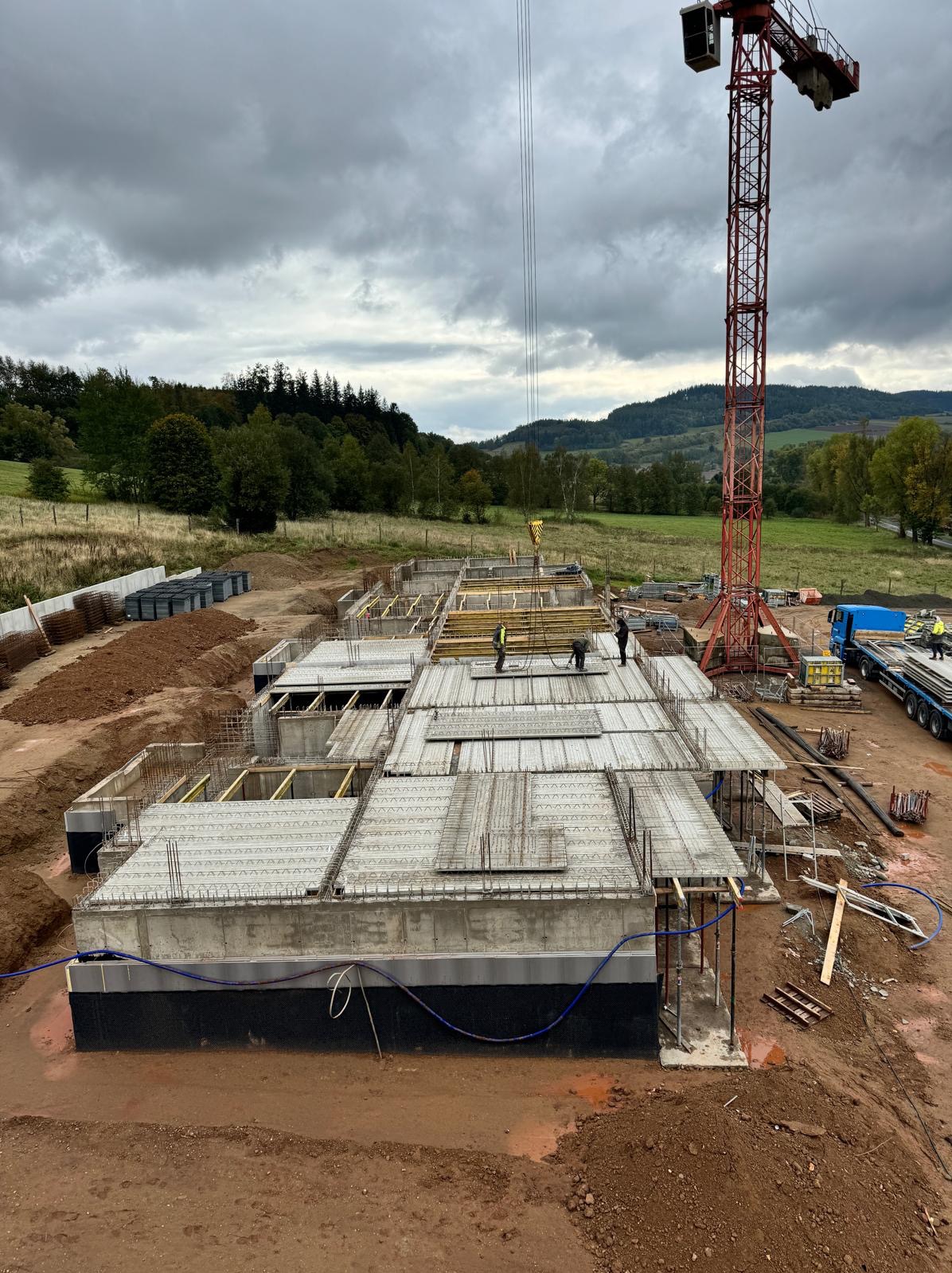 Budowa fundamentów domu z widoczną izolacją, zbrojeniami i elementami szalunkowymi. W tle krajobraz z lasem i wzgórzami, a także czerwony dźwig budowlany.