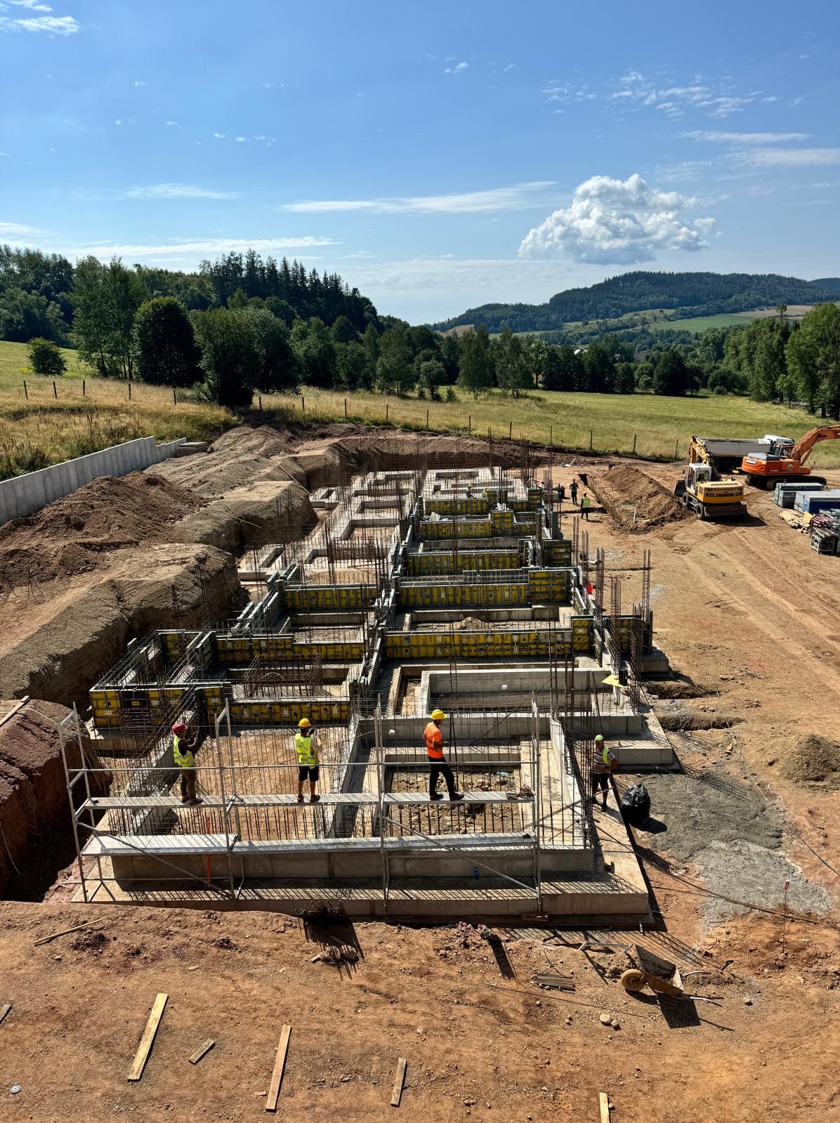 Budowa fundamentów domu jednorodzinnego z użyciem szalunków i zbrojenia, widok z góry na plac budowy z pracownikami w kaskach i maszynami budowlanymi w tle.