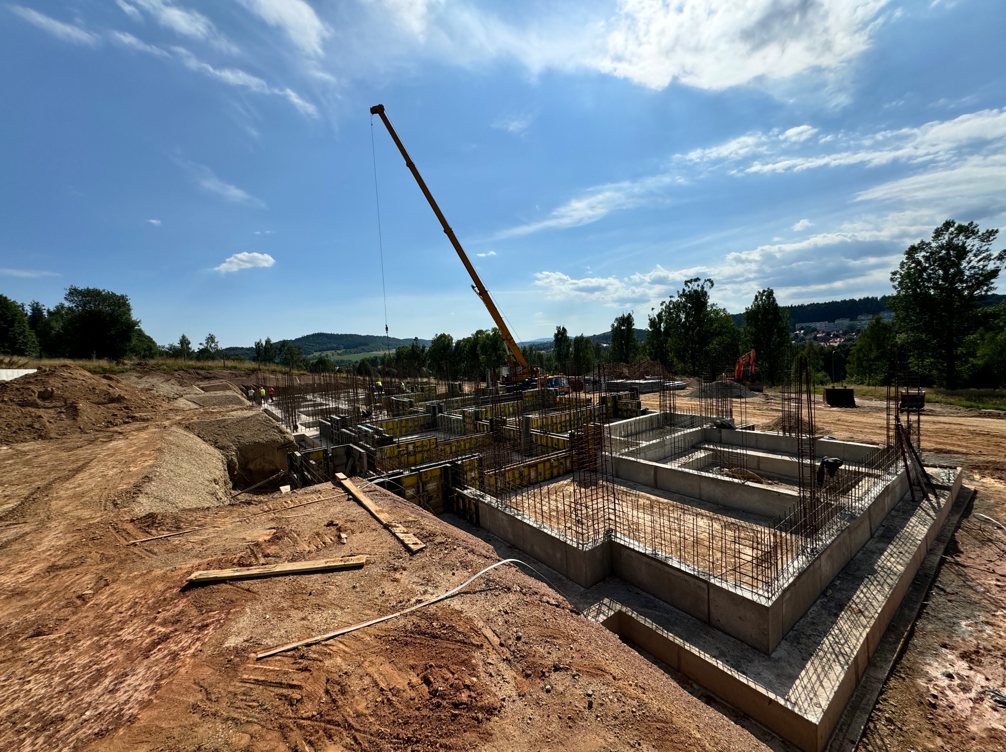 Szeroki kadr na fundamenty domu w trakcie budowy, z widocznym żurawiem budowlanym na tle błękitnego nieba z chmurami. Widoczne szalunki i zbrojenia.