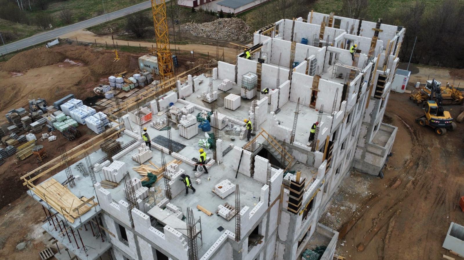 Wysoka perspektywa na budowę wielopiętrowego budynku z betonu komórkowego. Widoczni pracownicy w kaskach, zbrojenia, palety z materiałami i ciężki sprzęt budowlany.