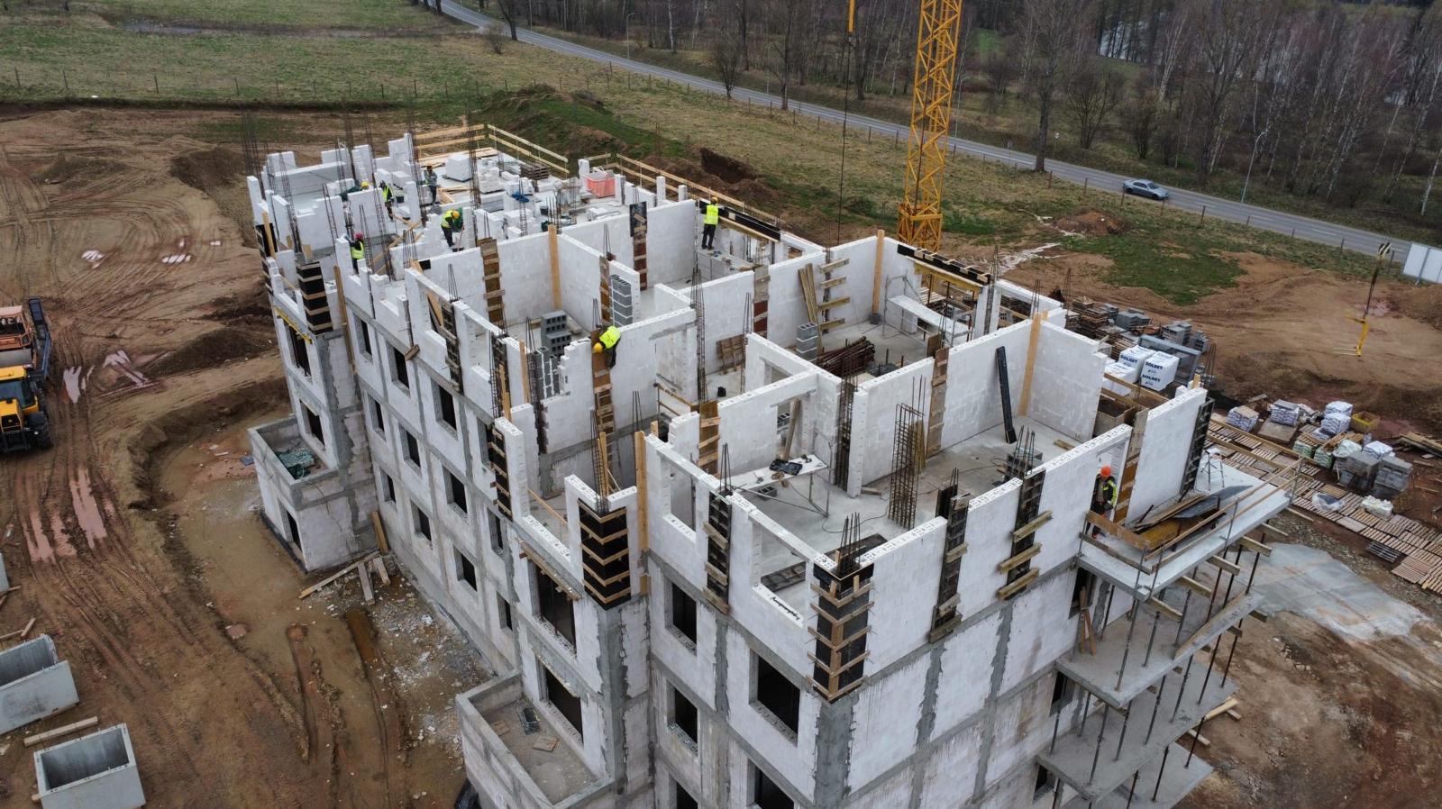 Wysoki budynek w trakcie budowy z betonu komórkowego, widok z góry. Żuraw budowlany w tle. Pracownicy w kamizelkach odblaskowych na budowie.