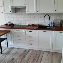 Biała kuchnia z drewnianym blatem i płytkami imitującymi cegłę, czarny kran i indukcja, drewniana podłoga i czarne uchwyty szafek.